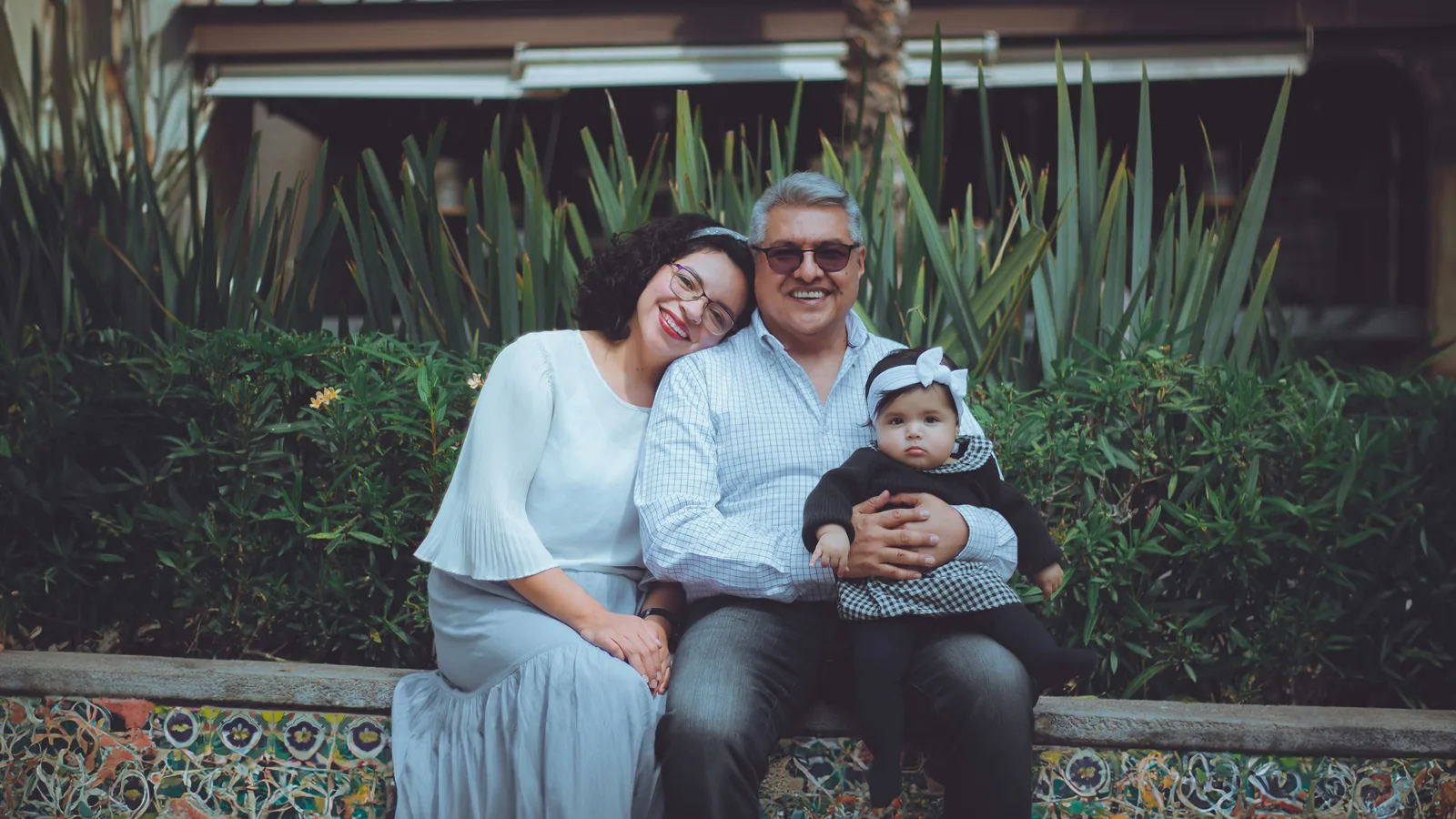 An older man, a young woman and a baby smiling together on a garden bench.